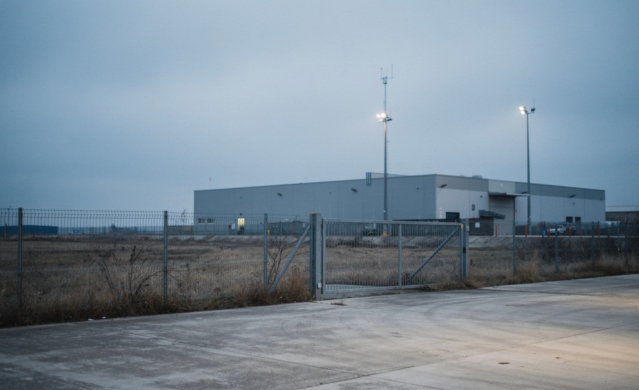A floodlit warehouse behind a perimeter fence at dusk